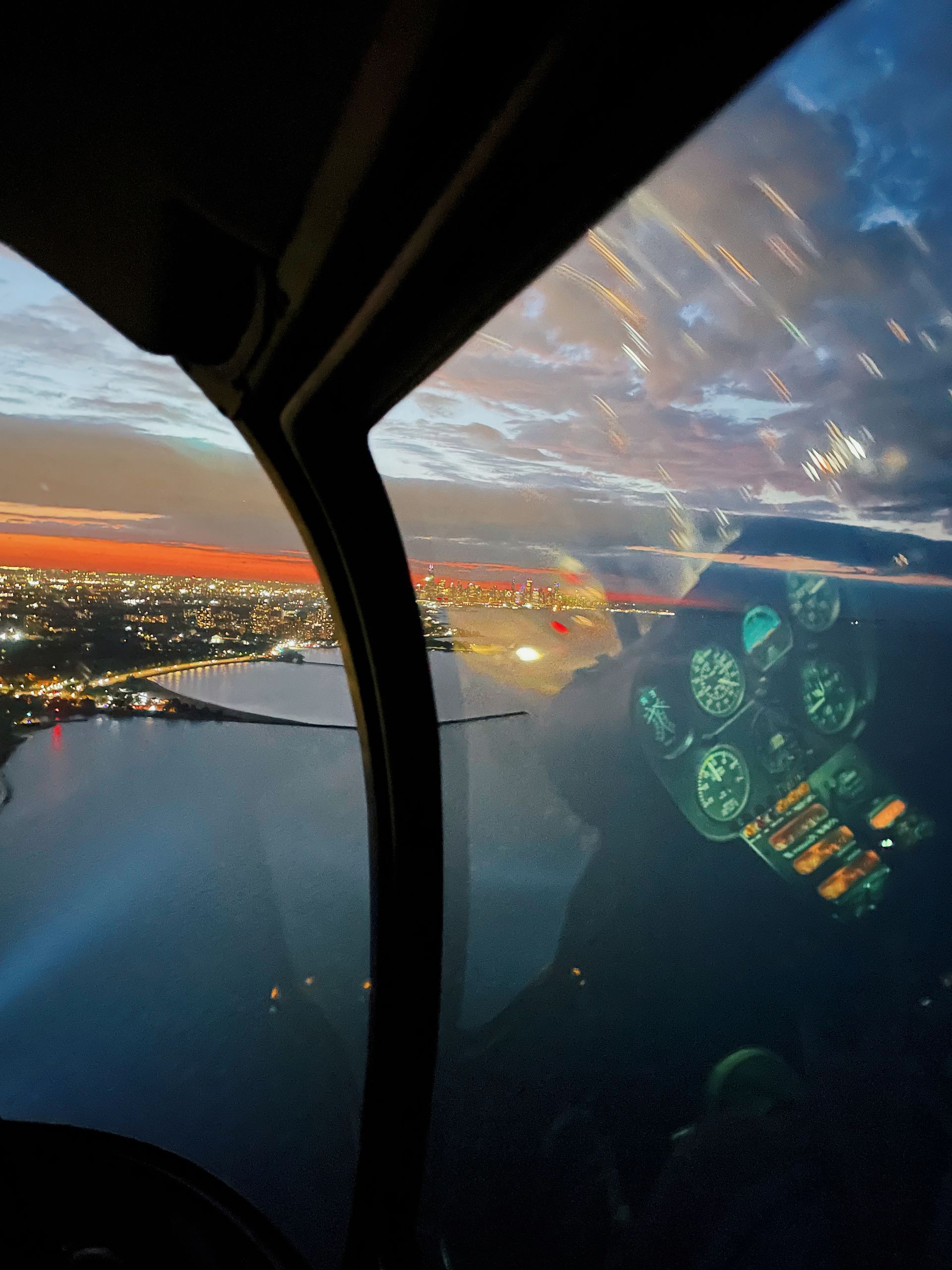 View from helicopter cockpit at dusk, overlooking city lights and water. Reflection of instruments visible on the window.