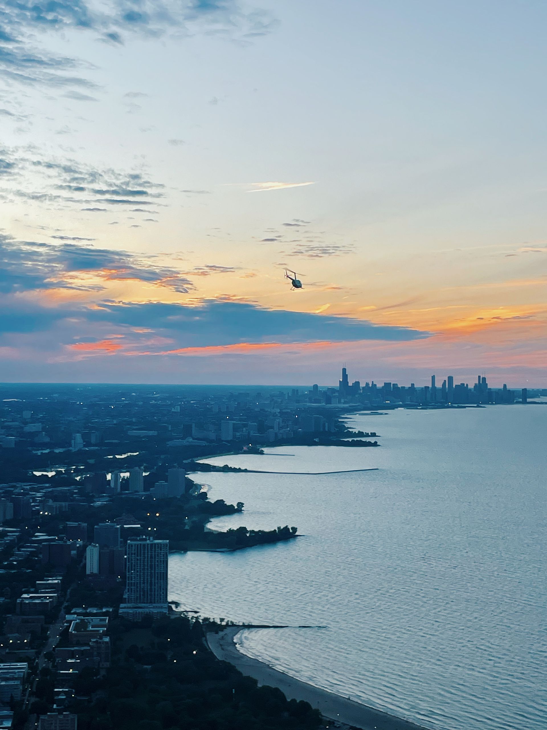 Coastal cityscape at sunset, with a helicopter flying in the sky over buildings and the ocean.