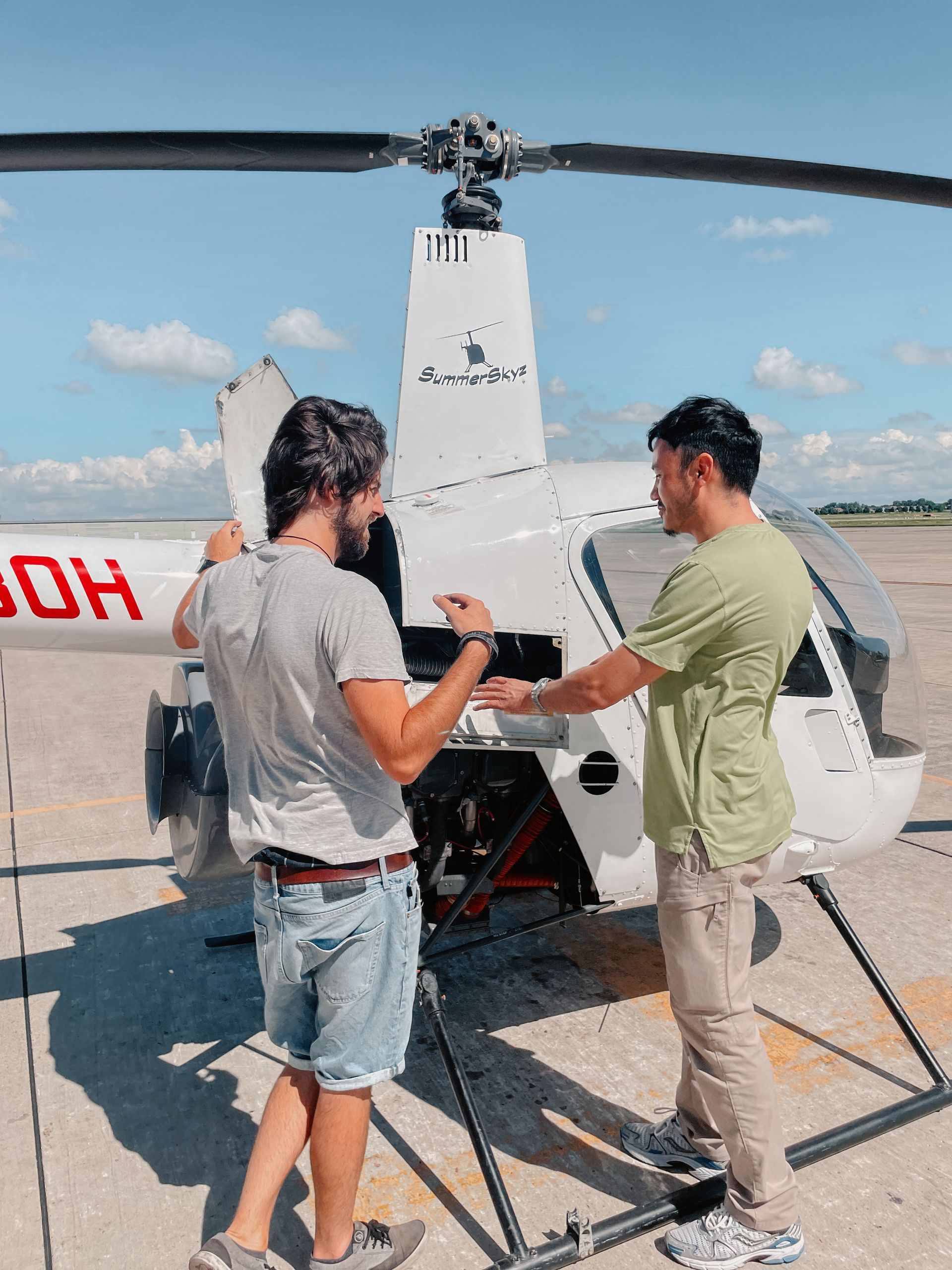 Two men inspecting a white helicopter outdoors on a sunny day; one points into the open compartment.