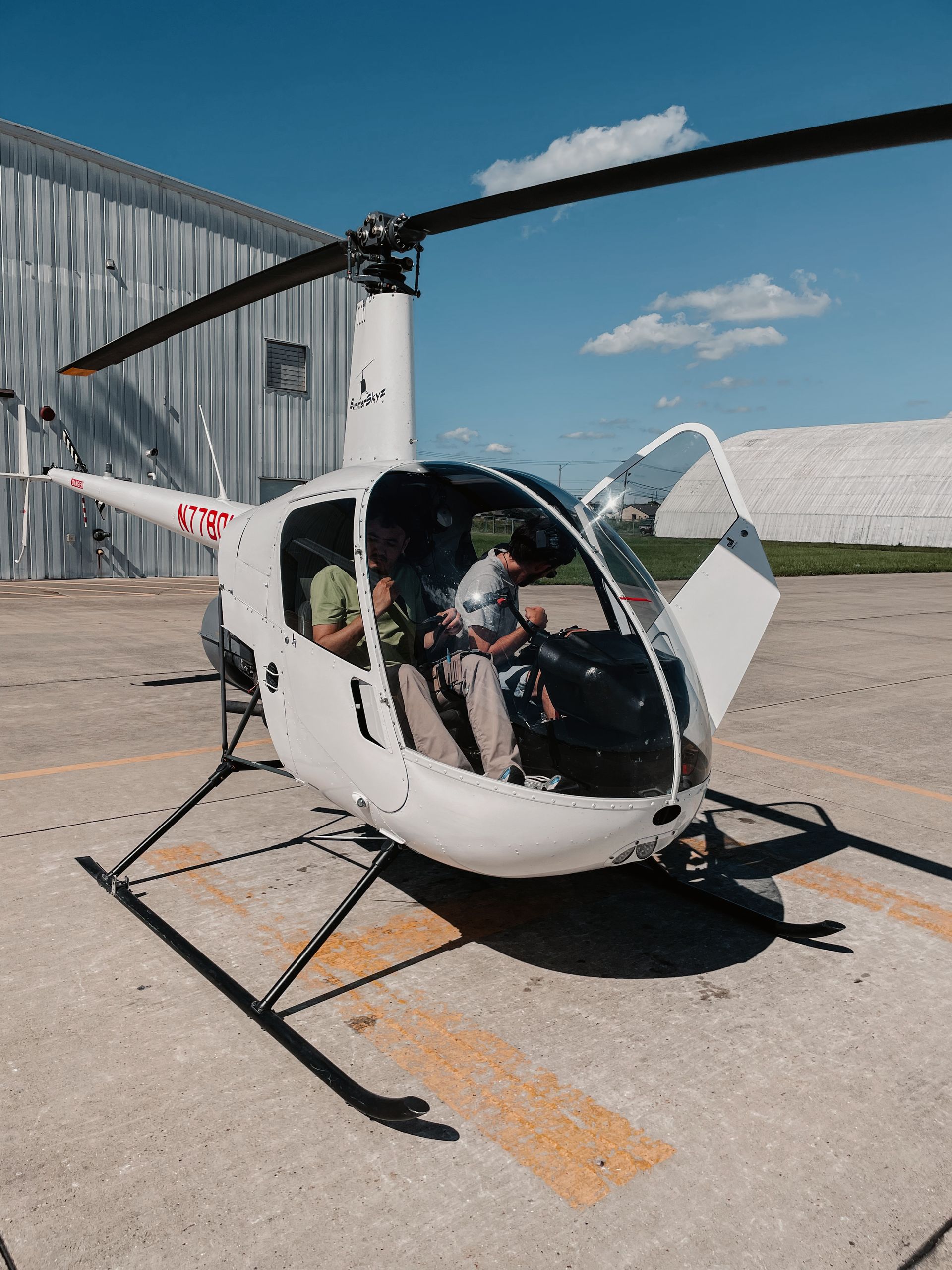 White helicopter on a tarmac; two people sit inside with the door open on a sunny day.