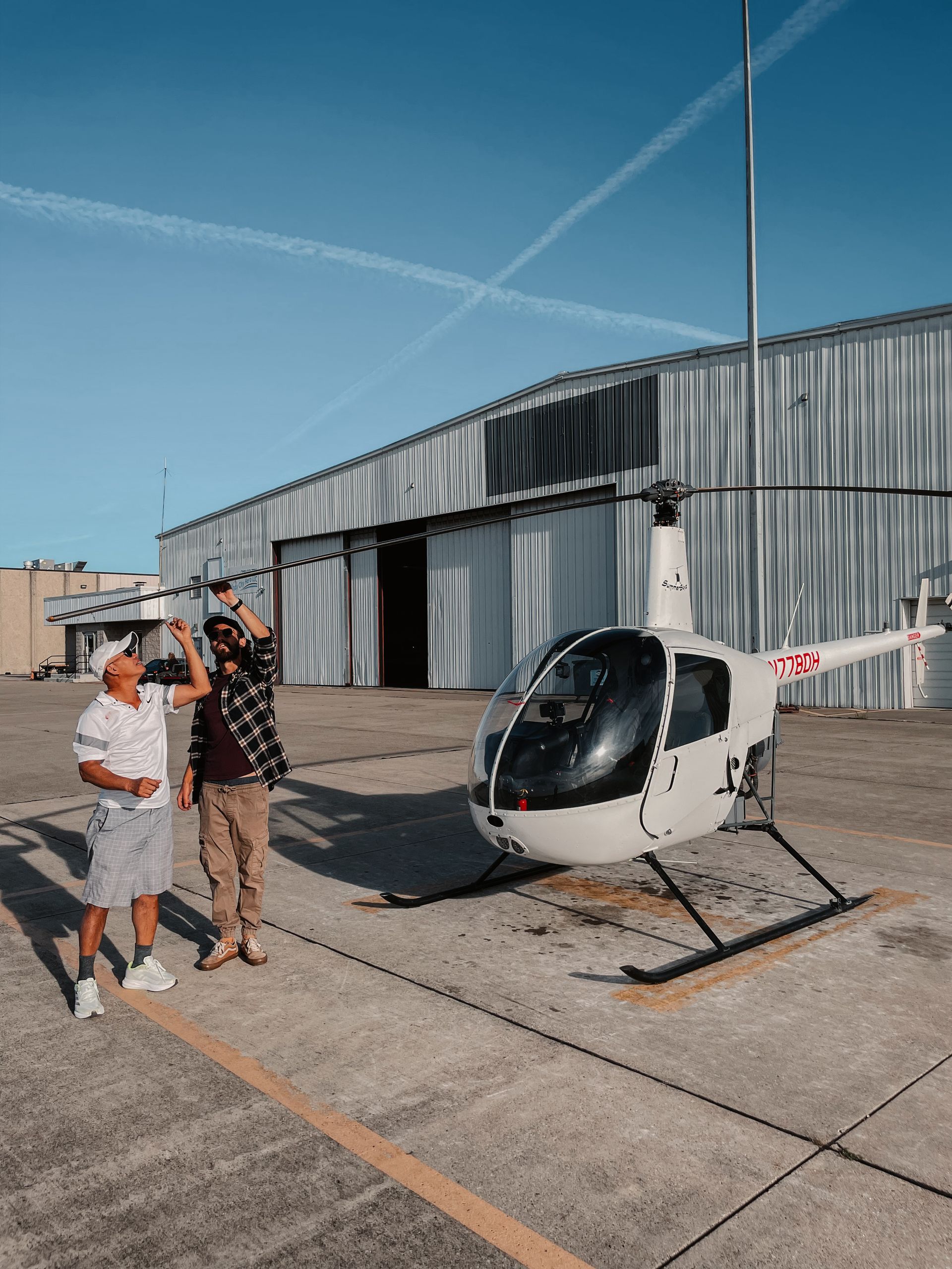 Two men by a white helicopter outside a hangar on a sunny day.