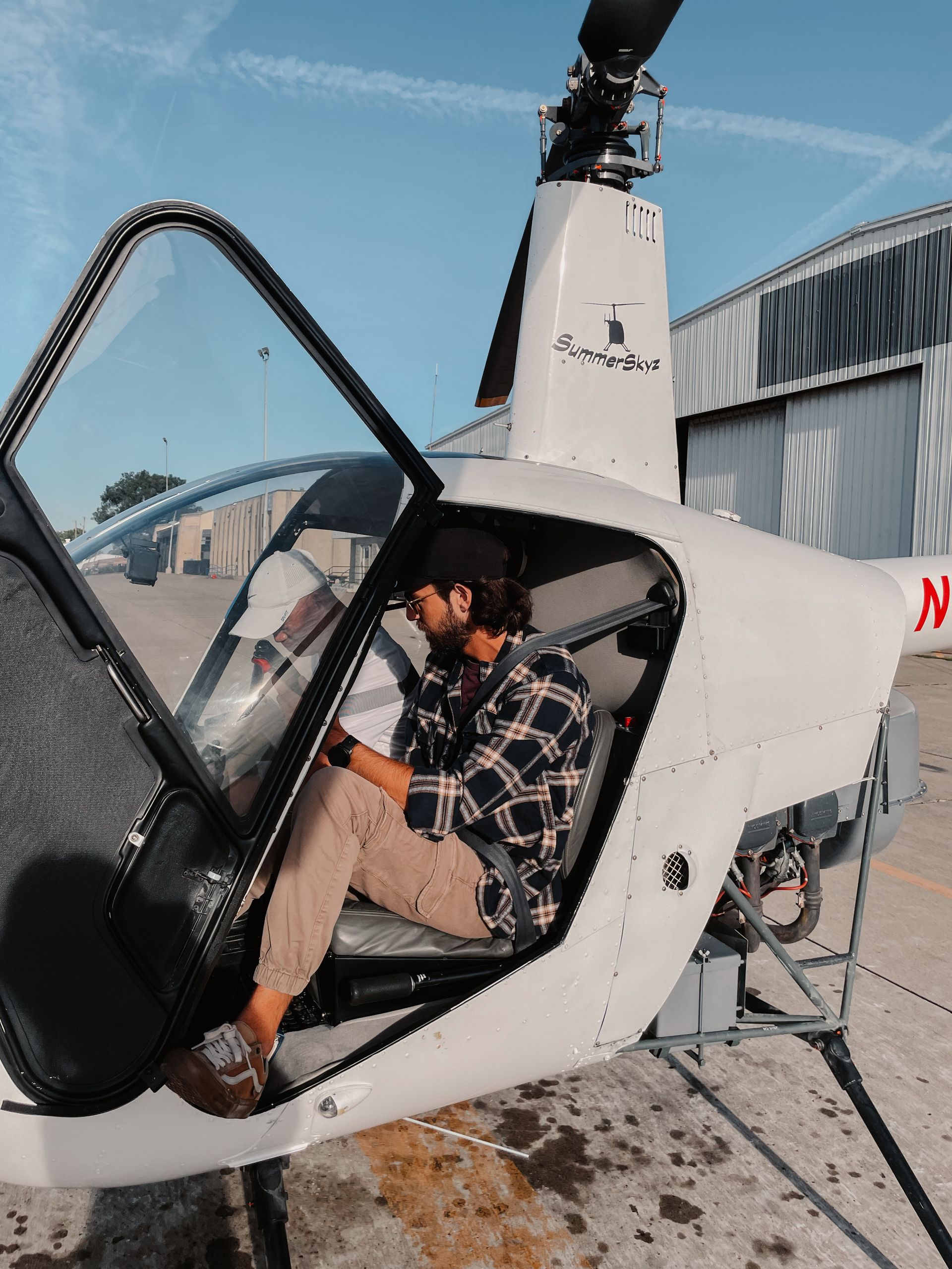 Man in a plaid shirt sits in a white helicopter cockpit with door open at an airfield.