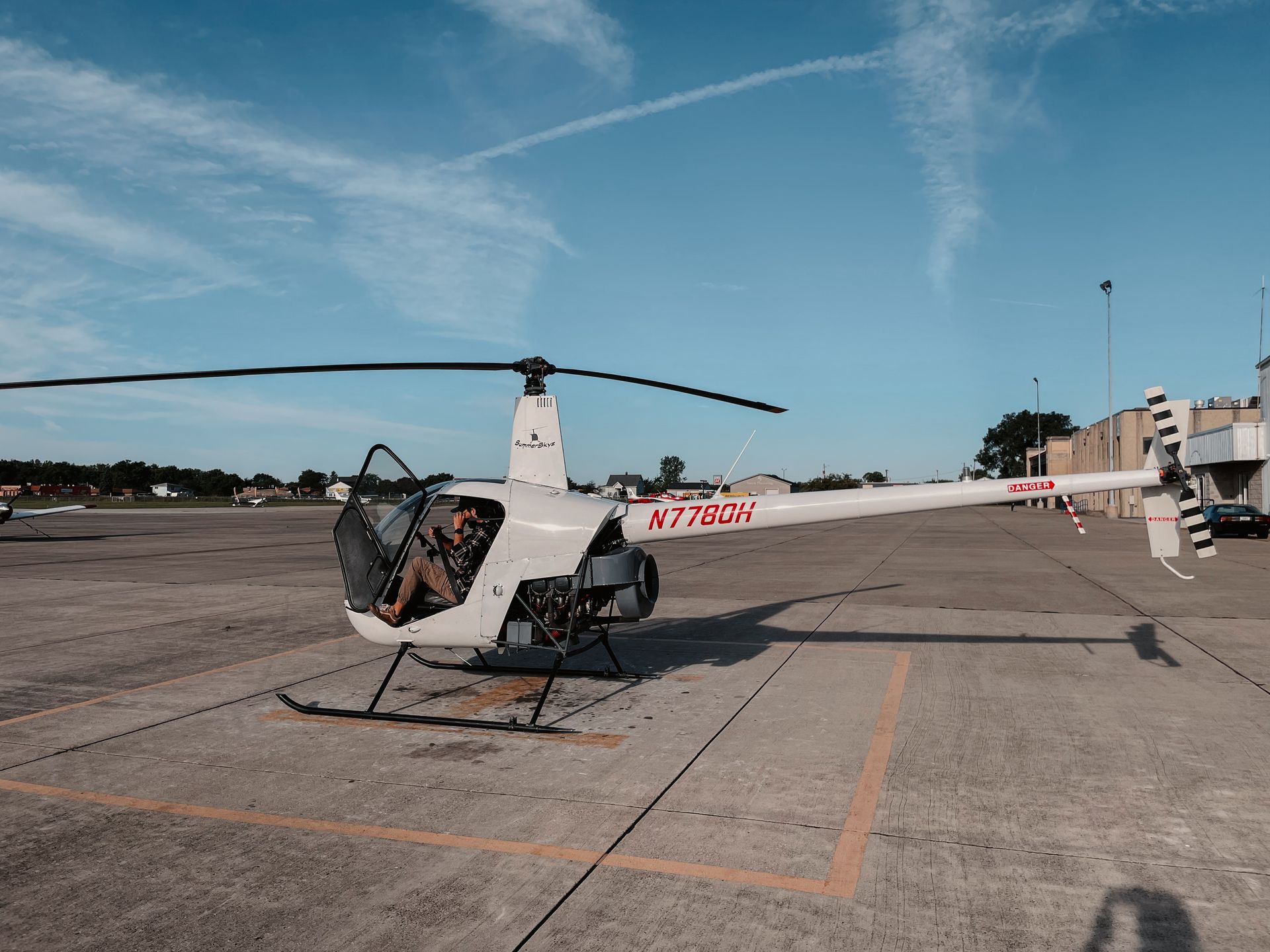 White helicopter on a tarmac, pilot visible in cockpit, blue sky with contrail.