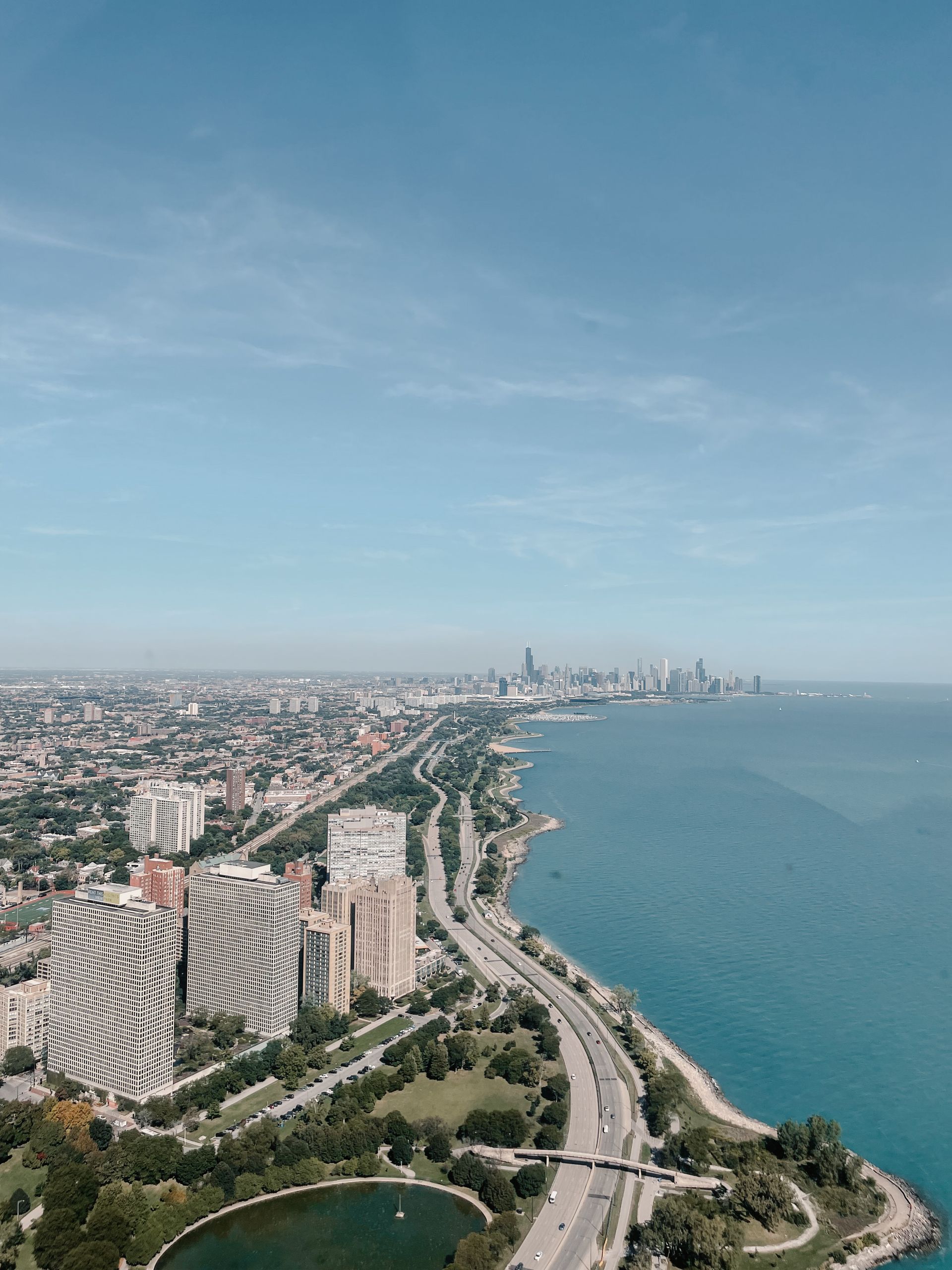 Aerial view of a city on the shore with tall buildings, highway, green park, and blue lake under a clear sky.