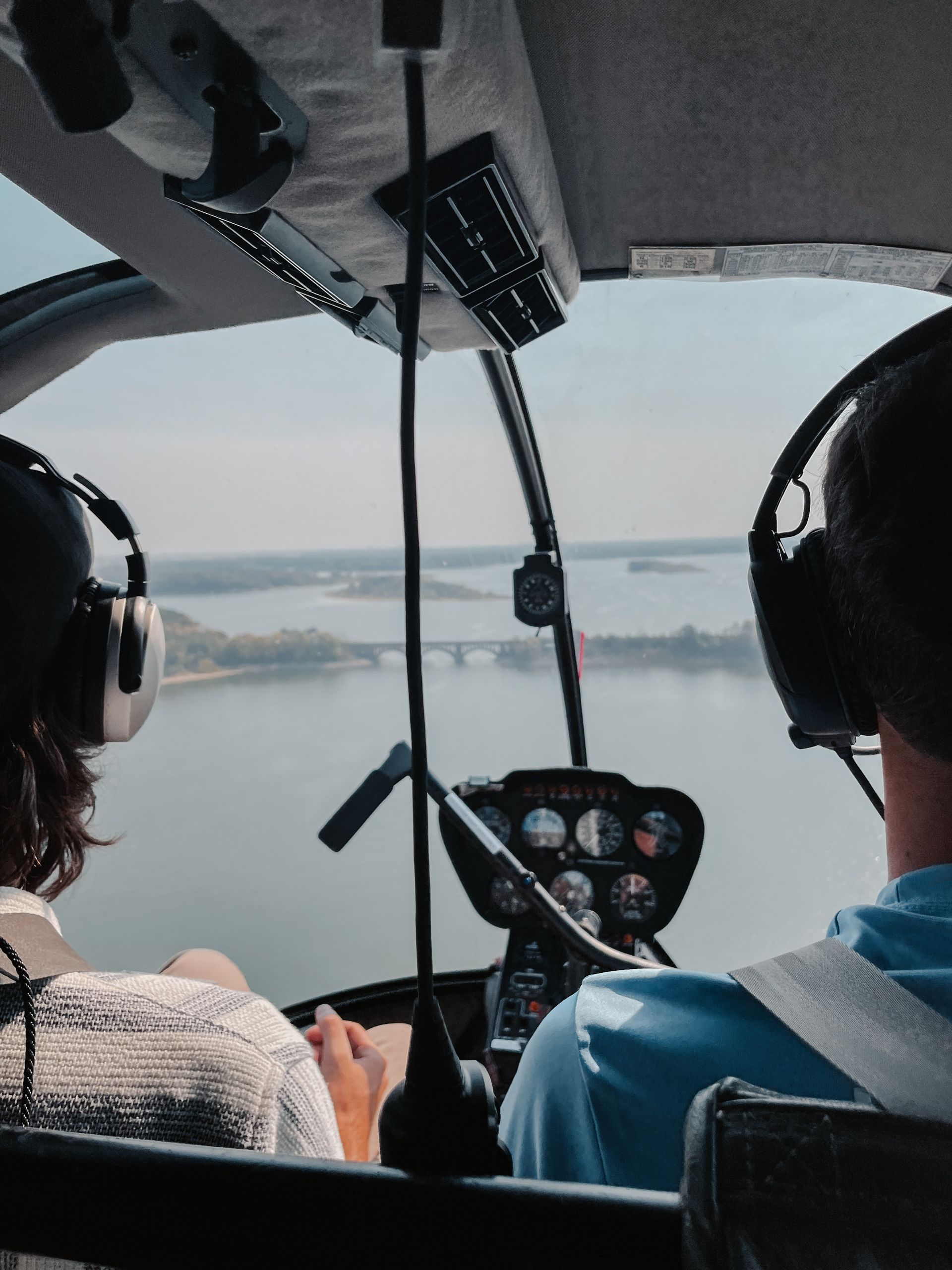 Inside a helicopter cockpit, two people fly over water toward a bridge.