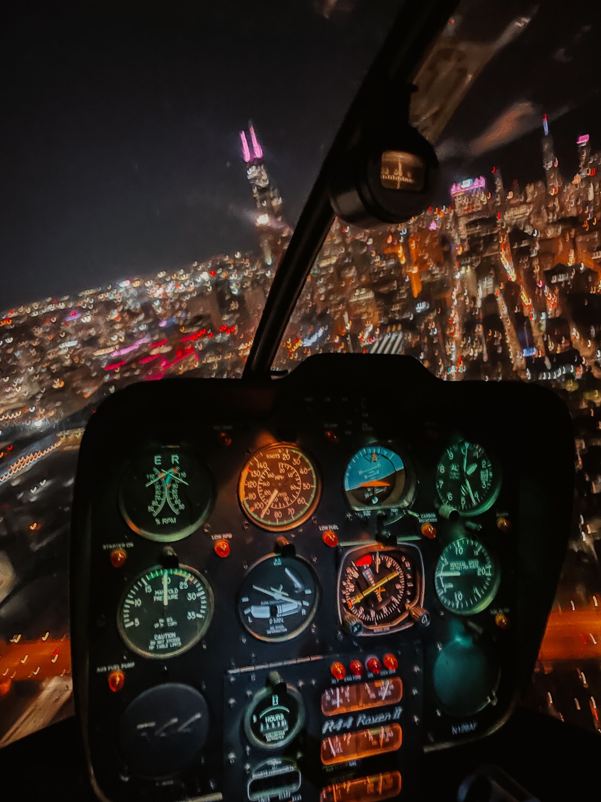 Helicopter cockpit view overlooking a brightly lit city skyline at night.