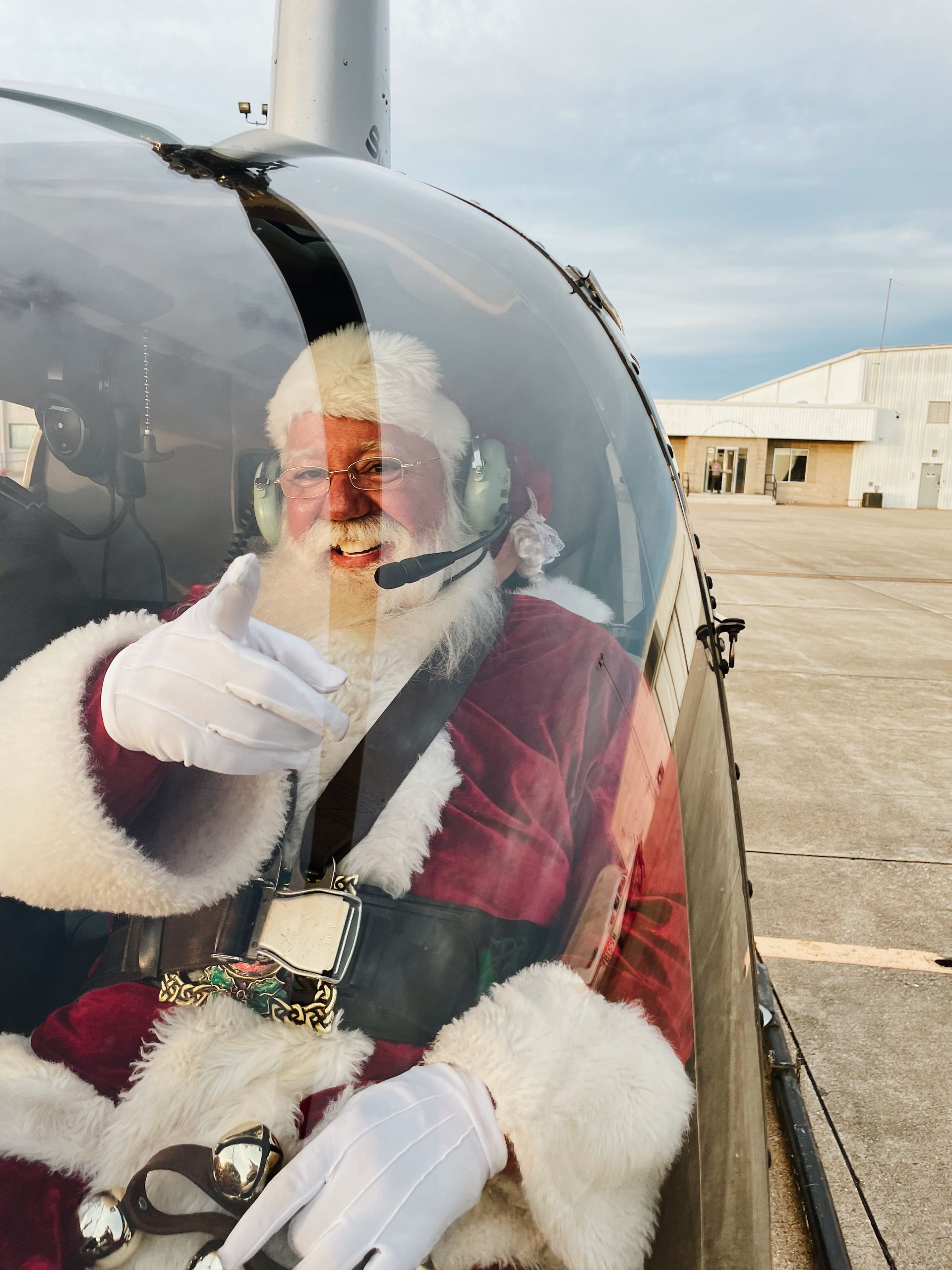 Santa Claus in a helicopter cockpit, giving a thumbs-up. Wearing a red suit, white gloves, and headset.