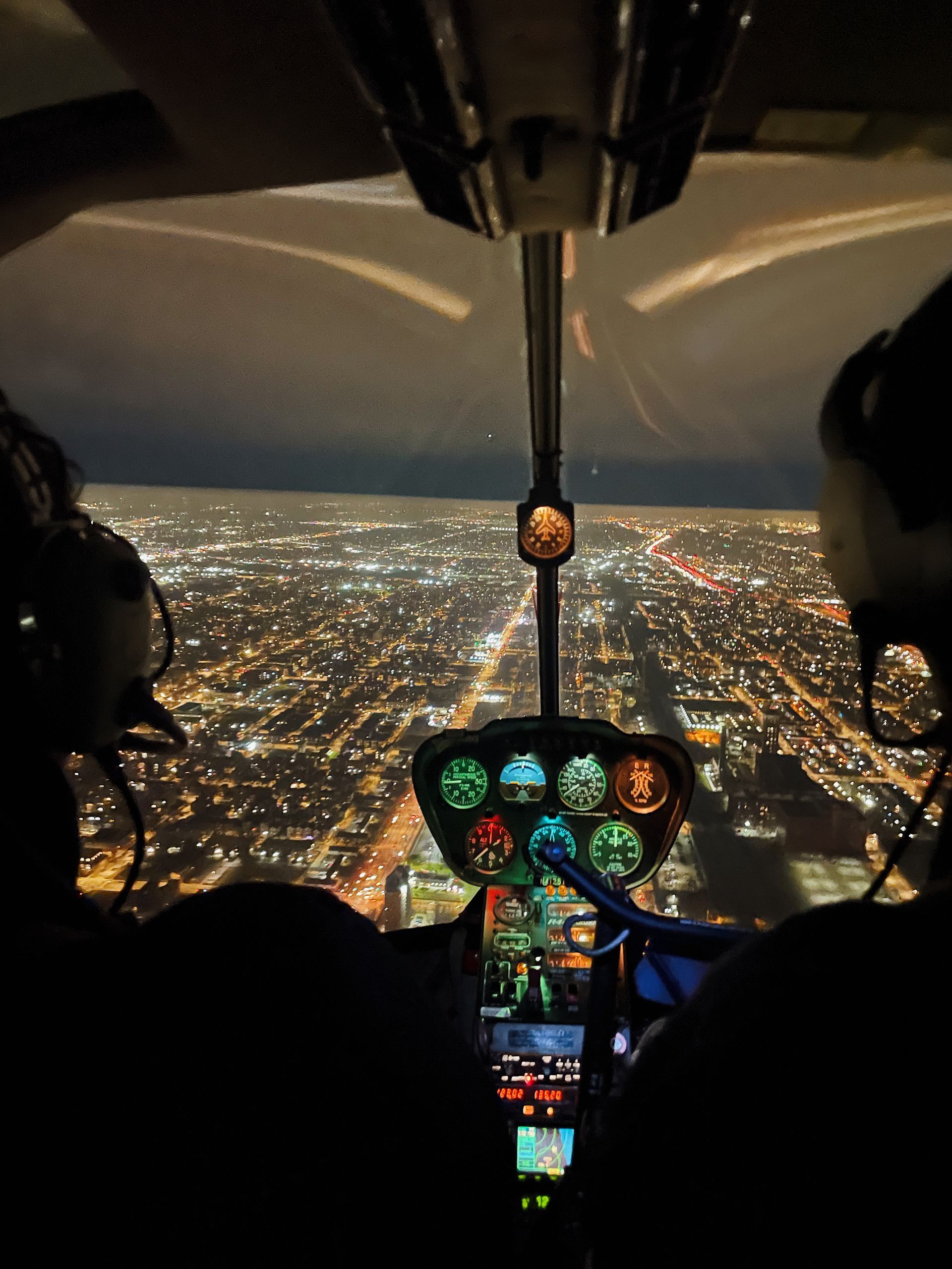Helicopter cockpit view at night over city lights. Gauges and pilot silhouettes visible.