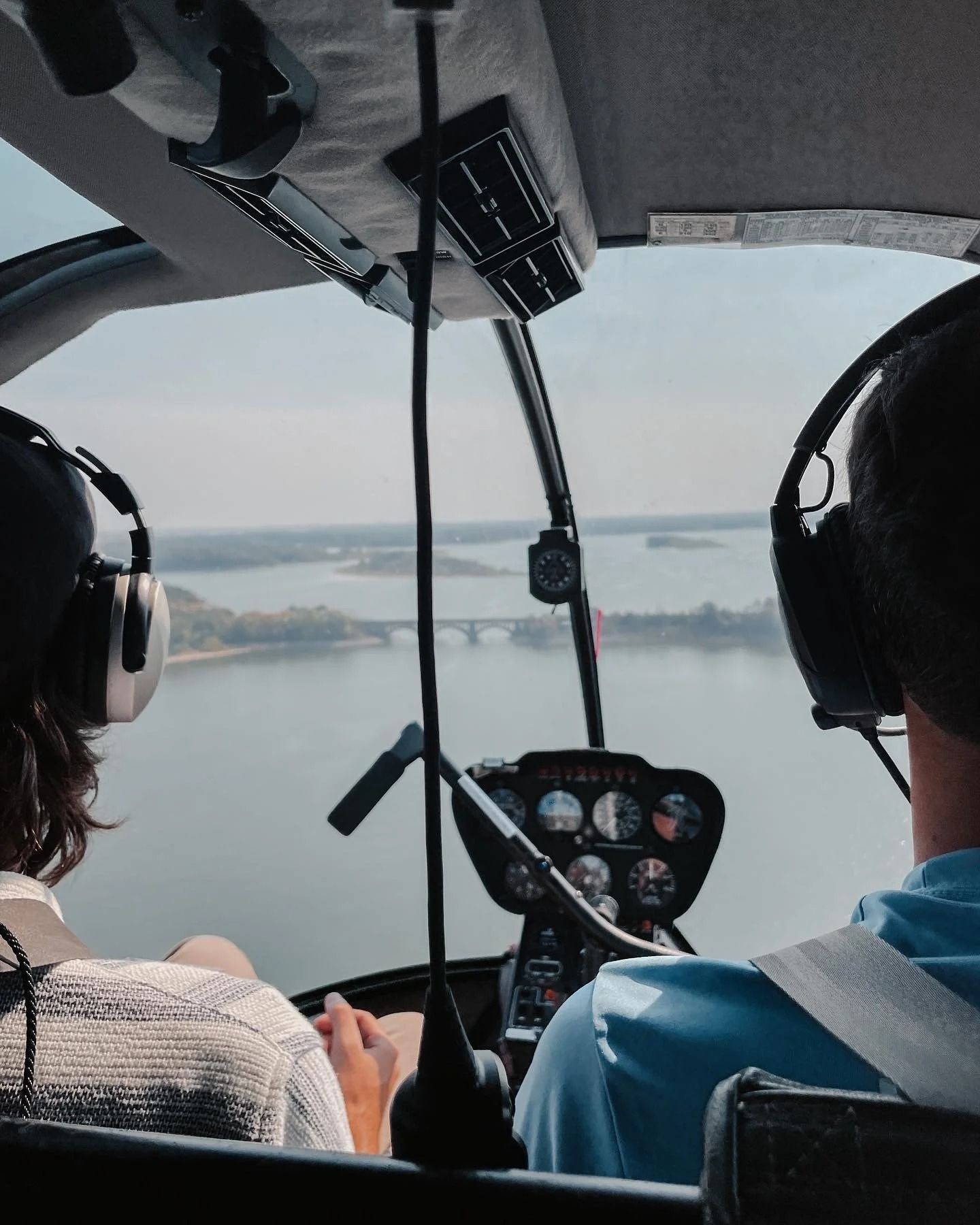 Inside a helicopter cockpit, two people look out over a body of water and a bridge.