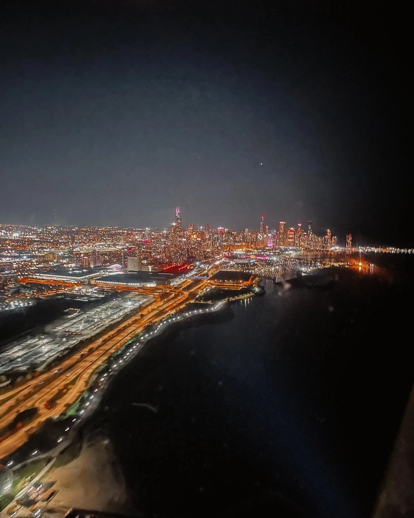 Night aerial view of a city skyline with glowing lights along the coast, a highway, and dark water.
