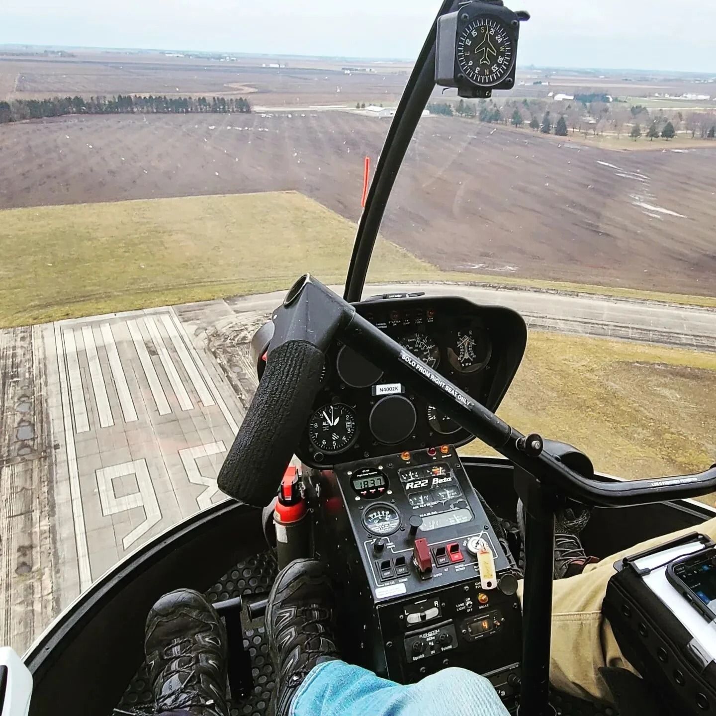 View from a helicopter cockpit looking over a runway. Pilot's feet on pedals, control stick and gauges visible.
