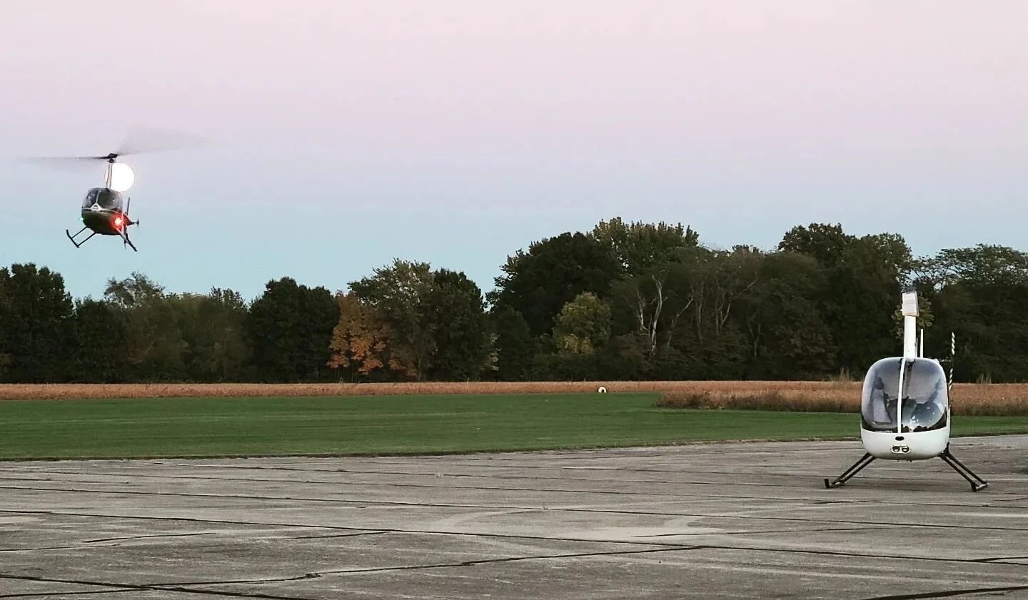 Two helicopters, one taking off, the other parked, on a concrete landing pad at dusk.