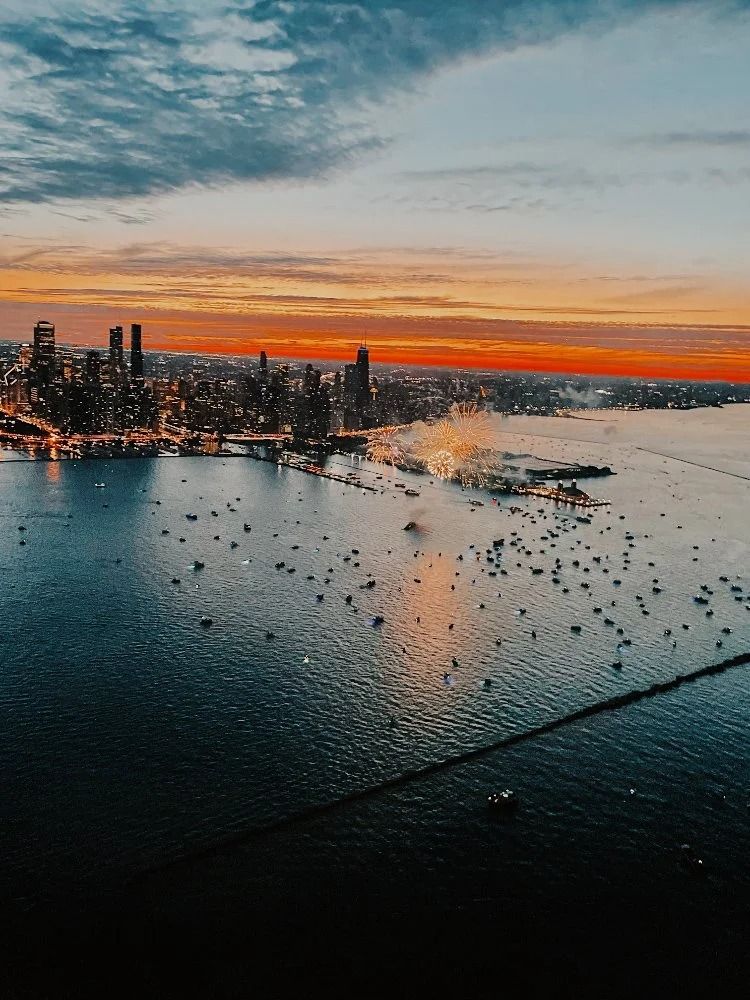 City skyline at sunset over a body of water dotted with boats; orange and blue hues.