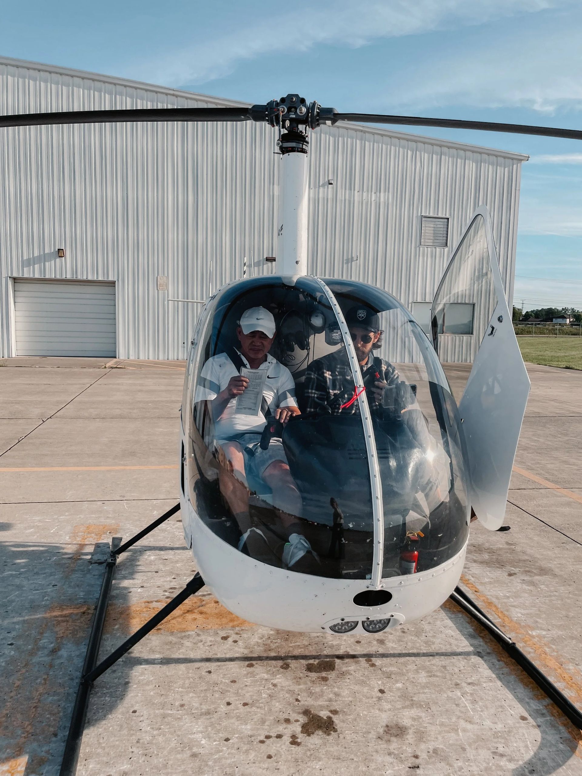 Two people in a white helicopter on a tarmac, hangar in the background. One gives a thumbs up.