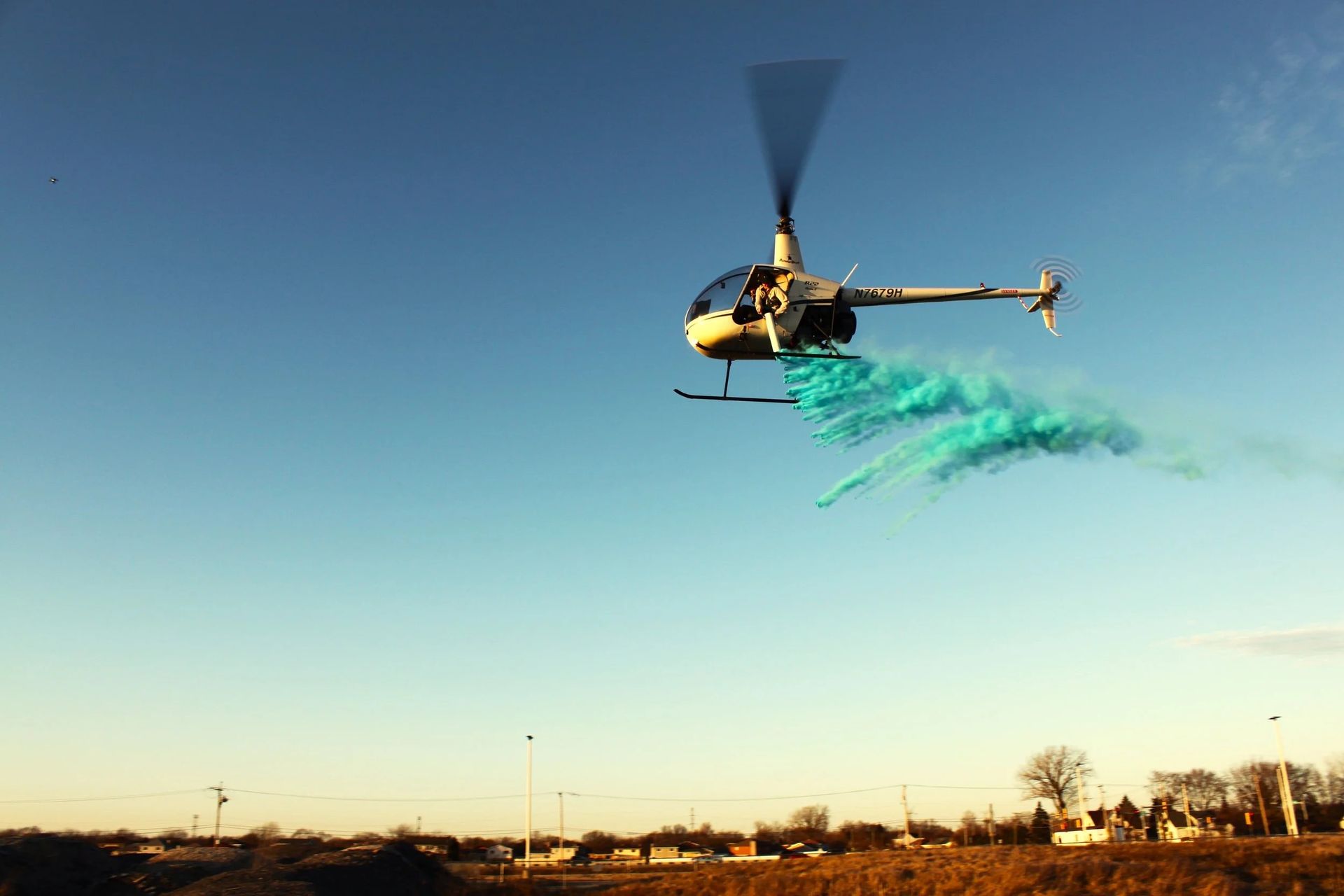Helicopter releasing blue powder, likely for a gender reveal, against a clear blue sky over a field.