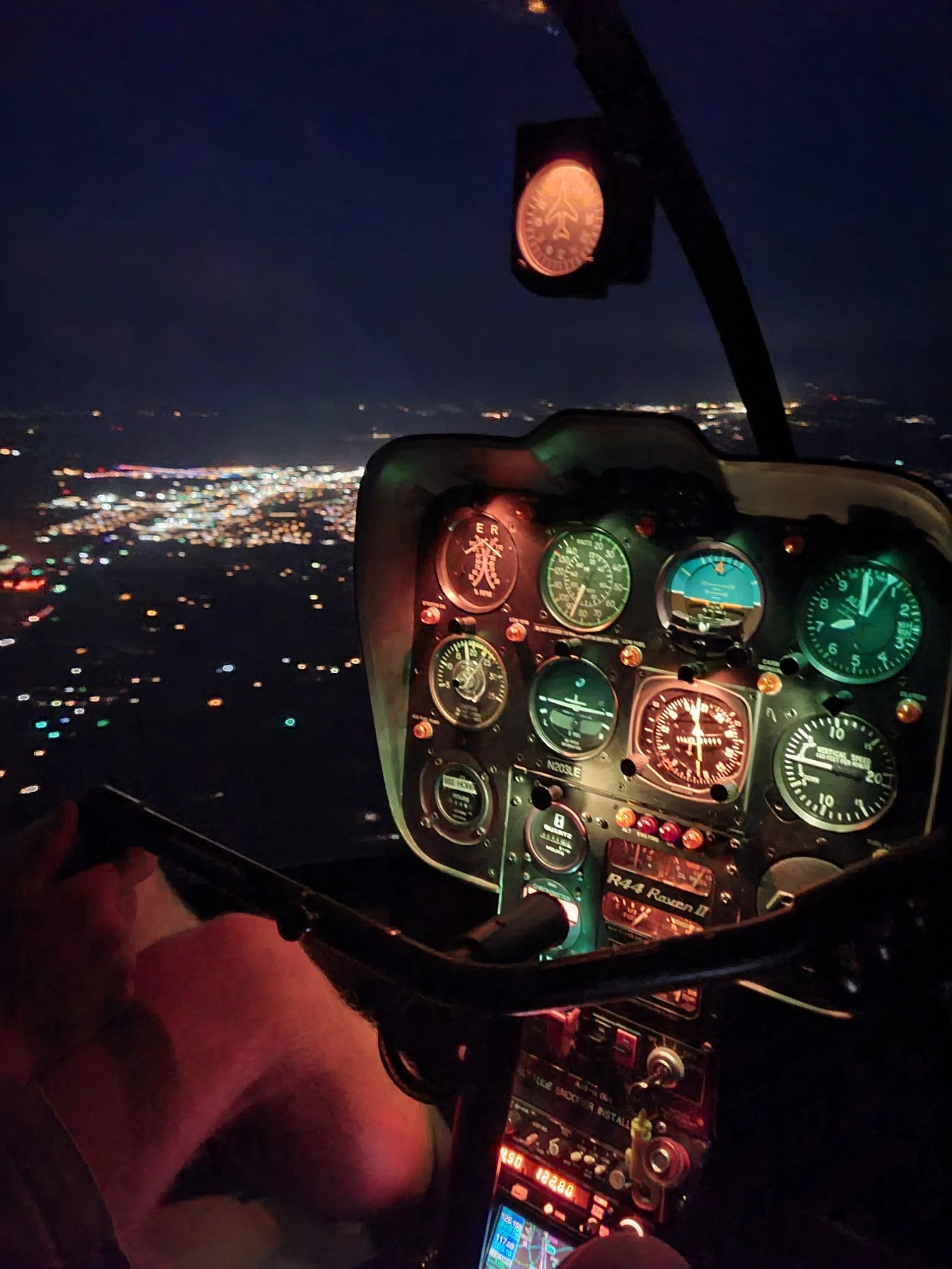 Night helicopter cockpit view with illuminated gauges and city lights below.
