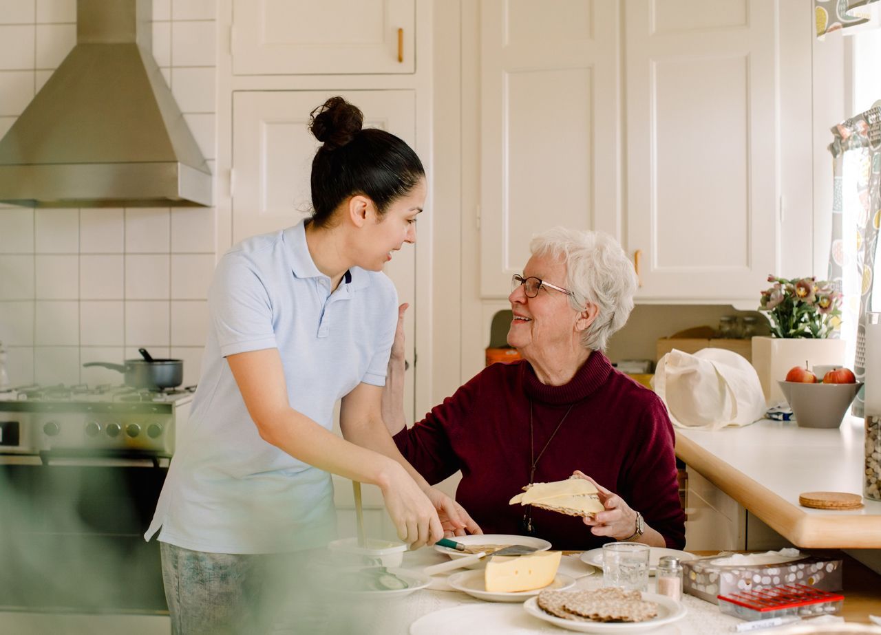 A caregiver in a light blue polo helps an older adult prepare a meal at a kitchen table.