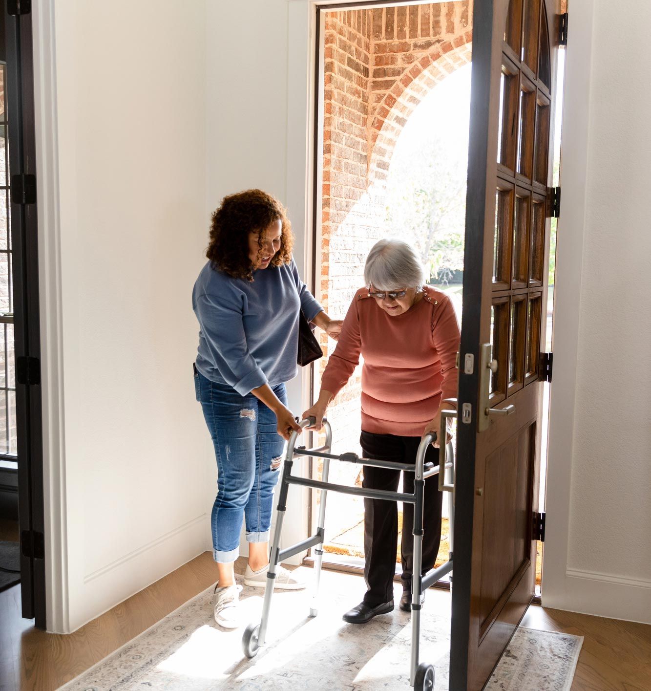 A caregiver supports an individual using a walker as they step through an open doorway into a home.