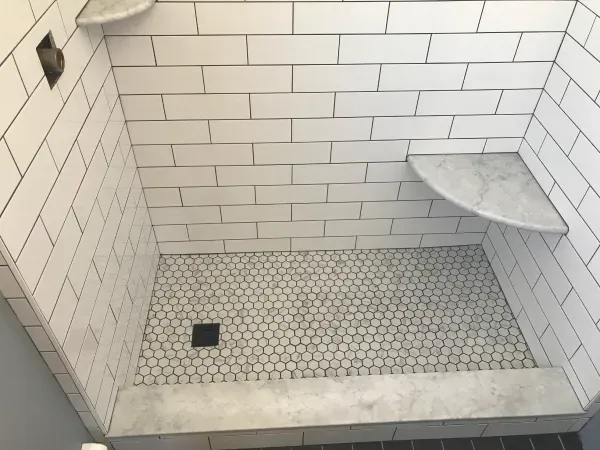 White tiled shower with a dark floor drain and built-in marble shelves.