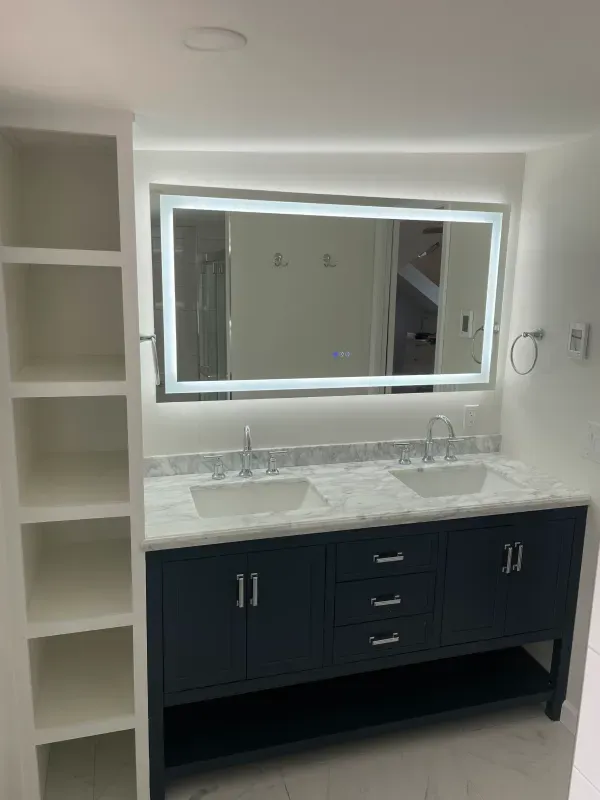 Modern bathroom with a dark blue double vanity, large lit mirror, and white shelves.
