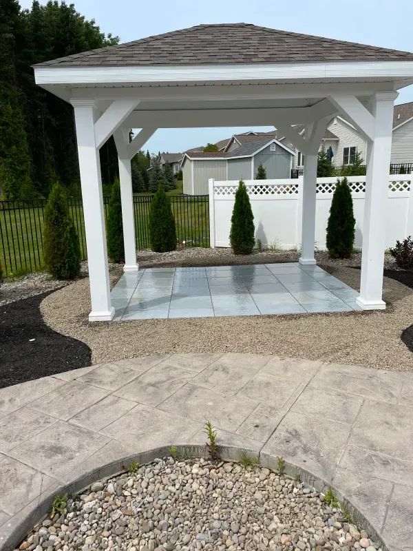 White gazebo with gray tiled floor in a backyard, surrounded by landscaping and a stamped concrete patio.
