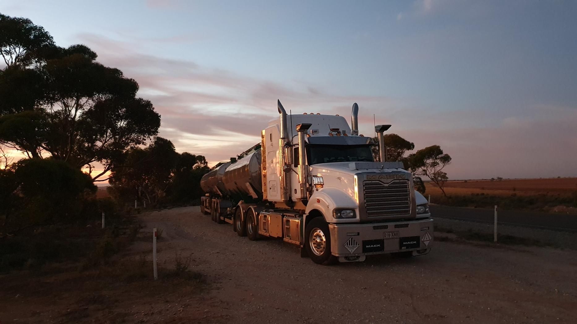 A Semi Truck Is Driving Down A Dirt Road At Sunset — Willow's Bitumen Haulage Pty Ltd In Northern Territory