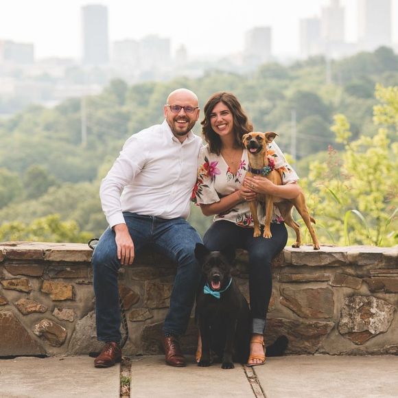 A man and a woman are sitting on a stone wall with their dogs.