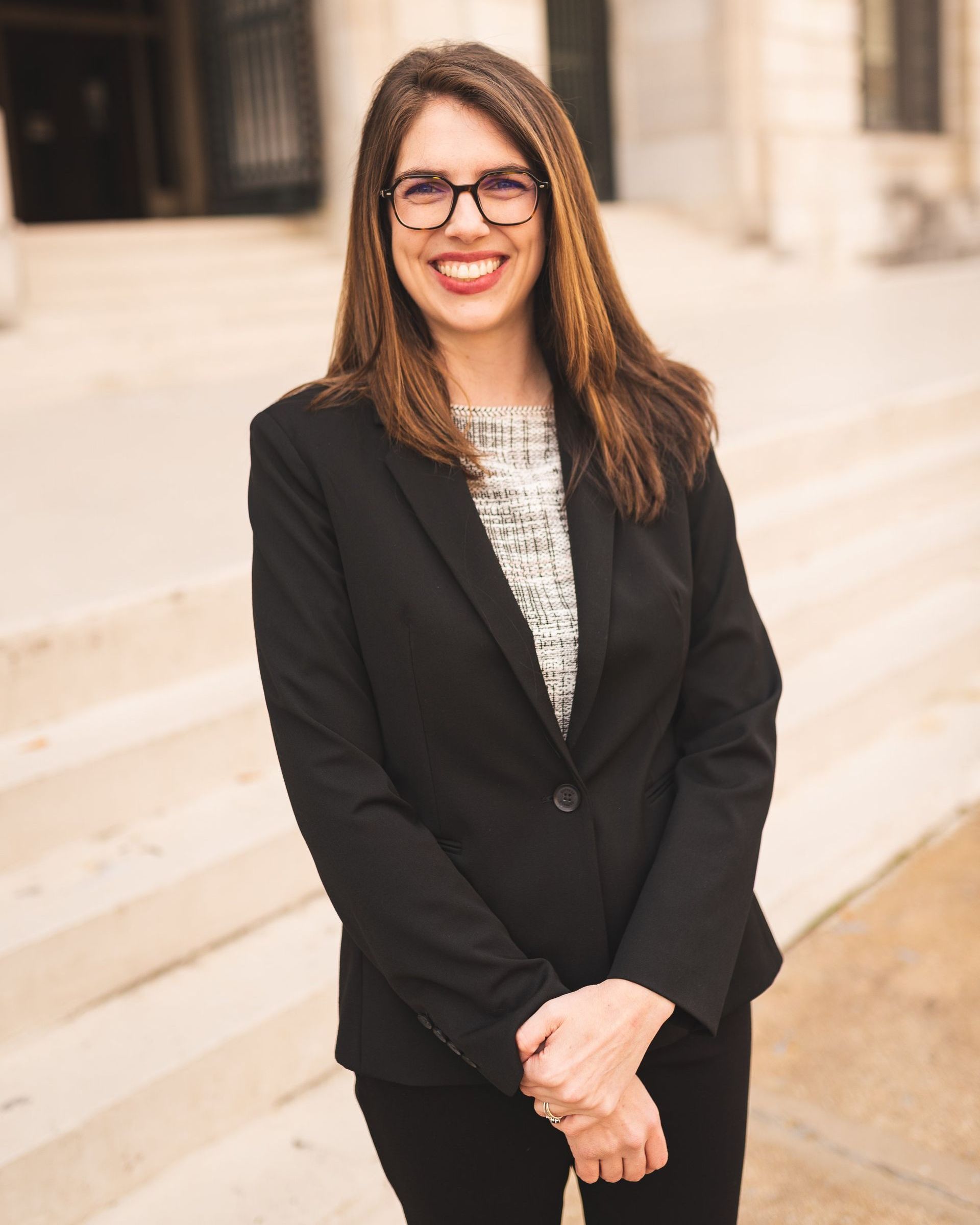 A woman in a black suit and glasses is standing in front of a building.