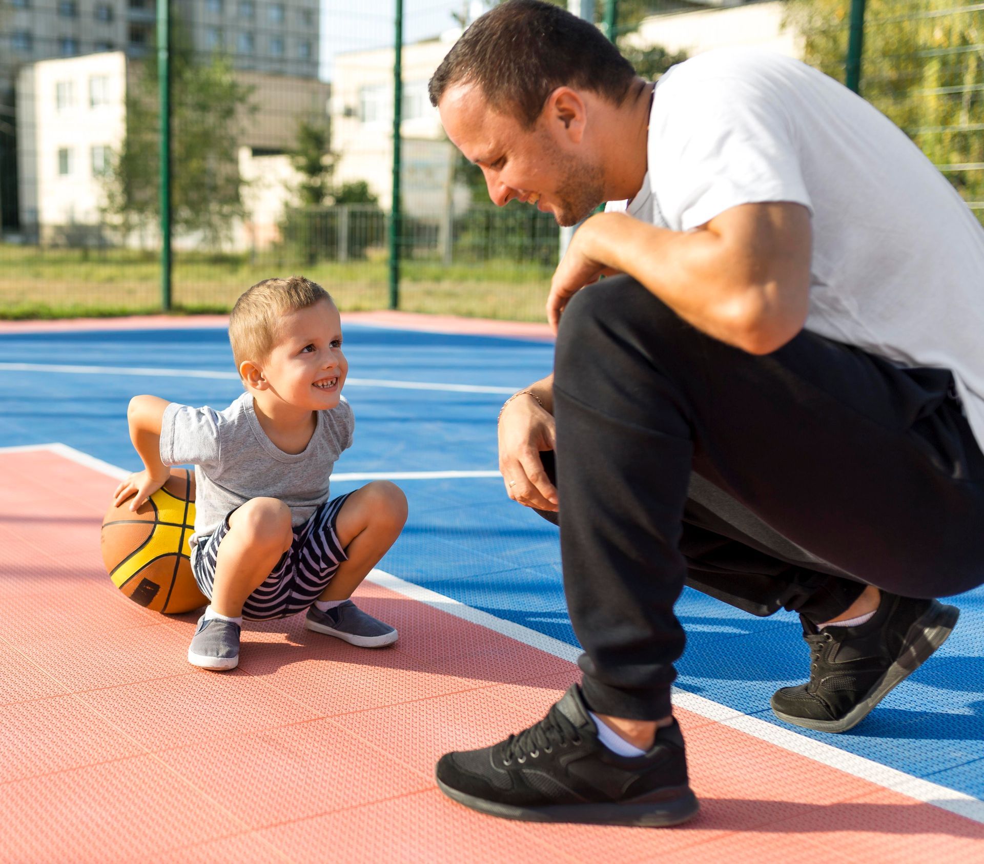 A man is squatting down next to a little boy sitting on a basketball court.