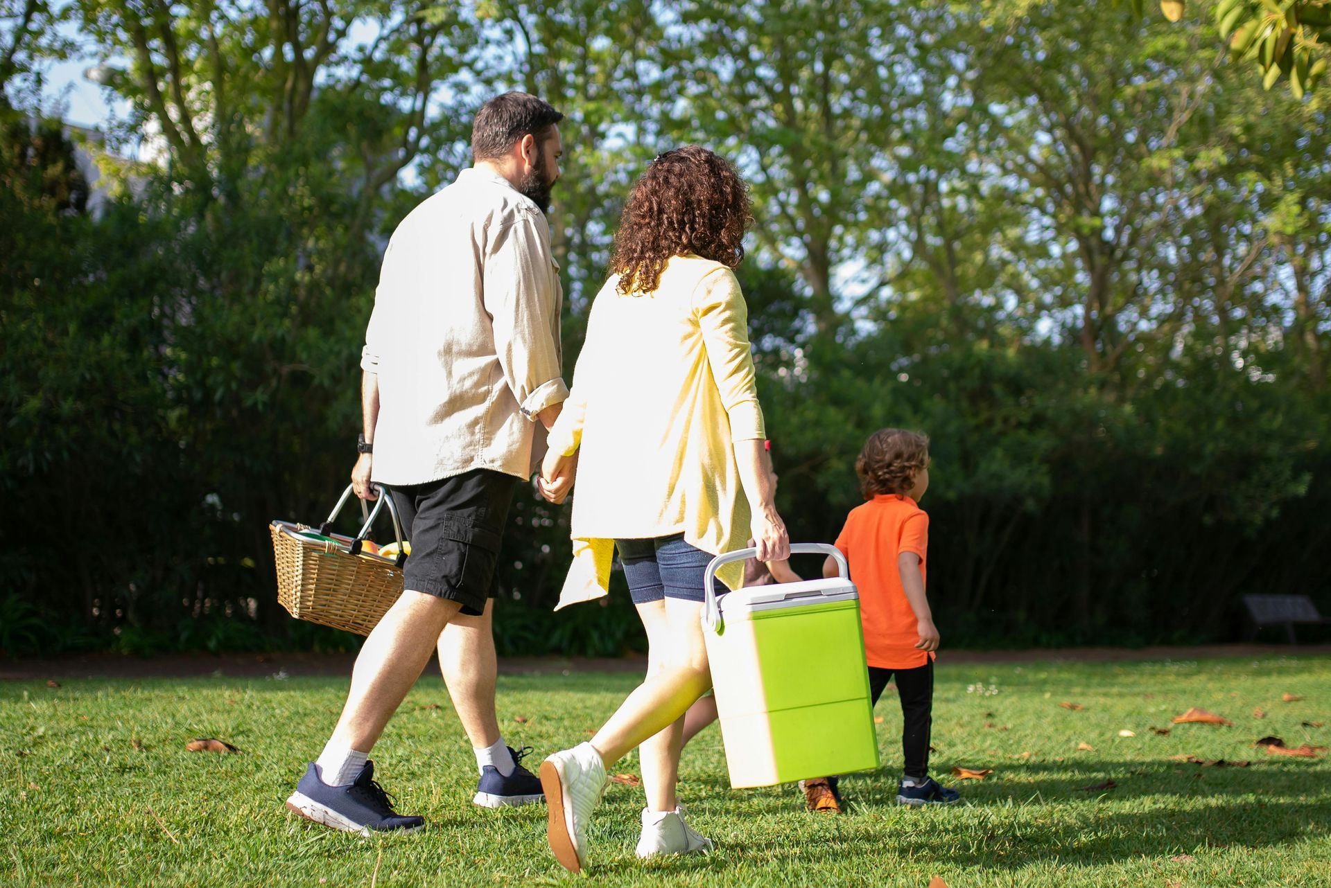 A family is walking through a park with a picnic basket and coolers.