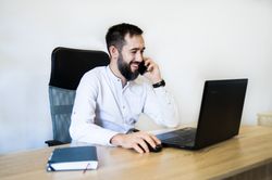Een man zit aan een bureau en glimlacht terwijl hij aan de telefoon praat naast een open laptop.