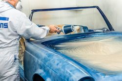Iemand spuit een blauwe autocarrosserie in een werkplaats, waarbij de voorruit is afgeplakt.