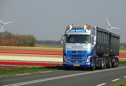 Een blauwe vrachtwagen rijdt over een landweg langs kleurrijke bloemenvelden en windmolens.
