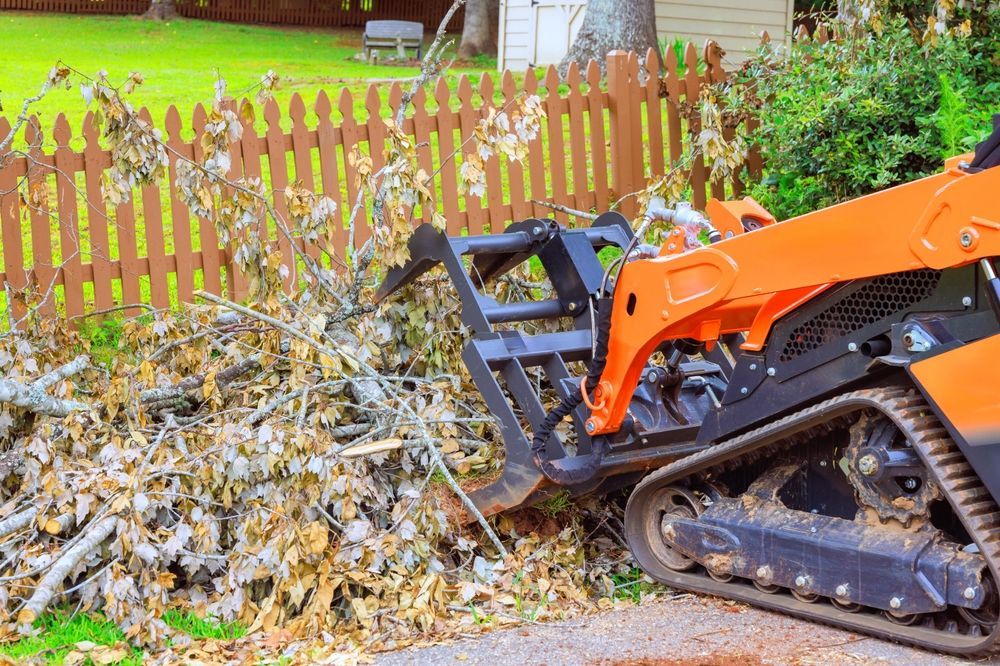 Orange mini loader pushing a large pile of yard debris beside a wooden fence