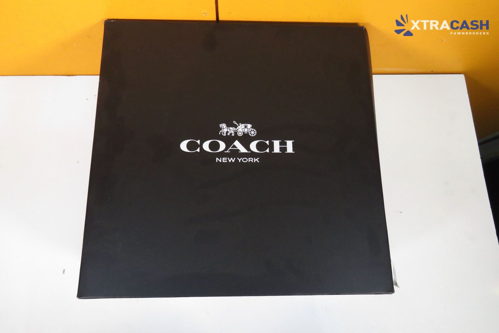 Black gift bag with white handles and matching box on a white surface against an orange background.