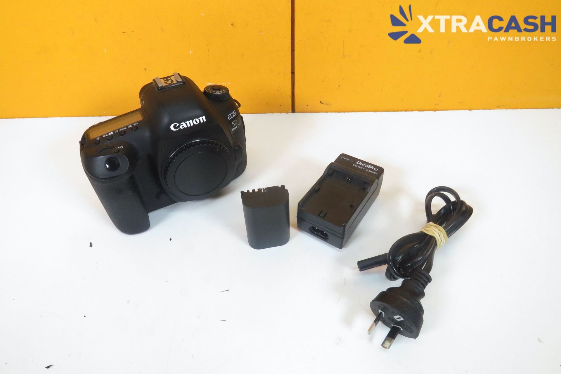 Canon camera with battery, charger, and power cord on a white surface under an Xtracash backdrop