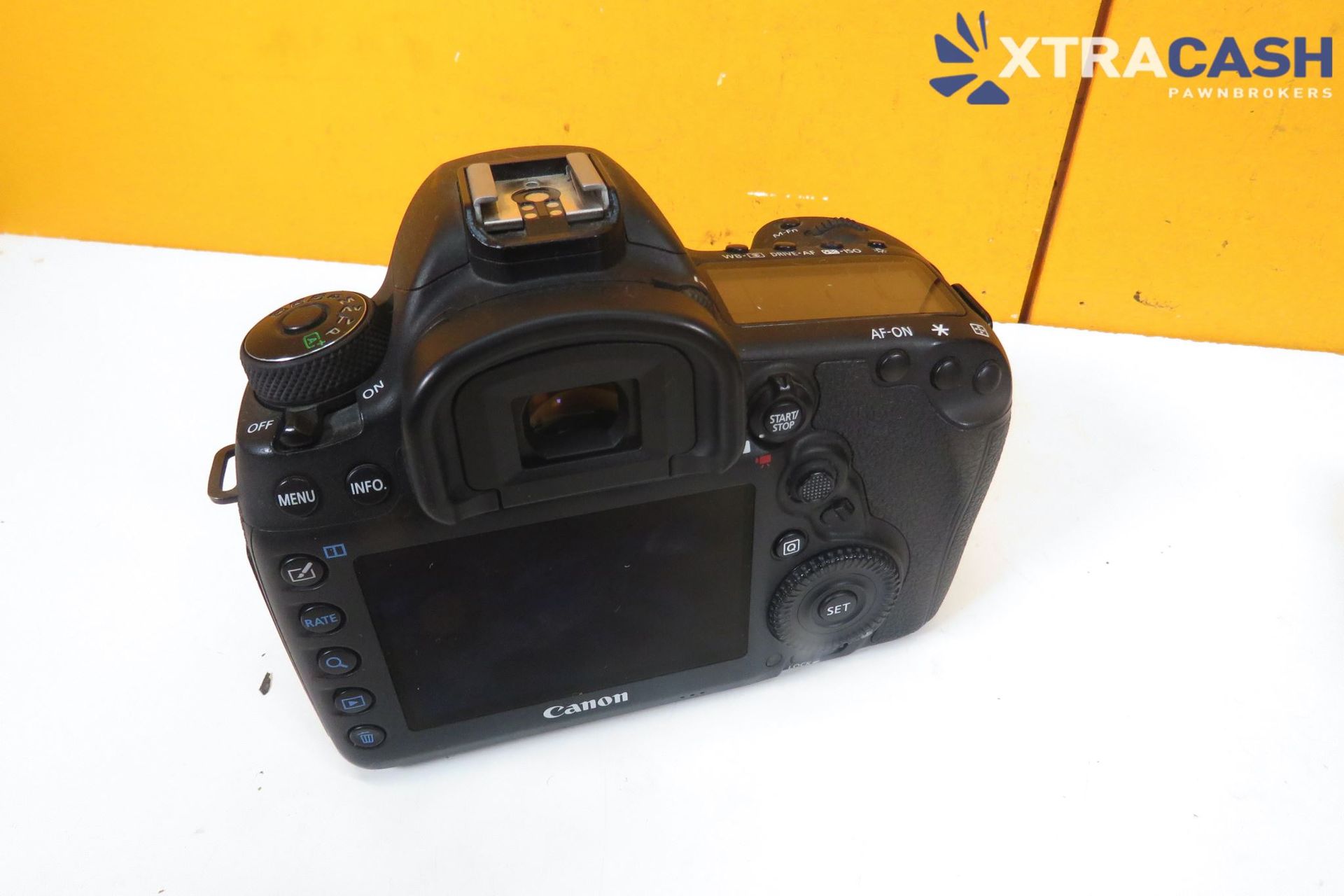 Canon camera with battery, charger, and power cord on a white surface under an Xtracash backdrop