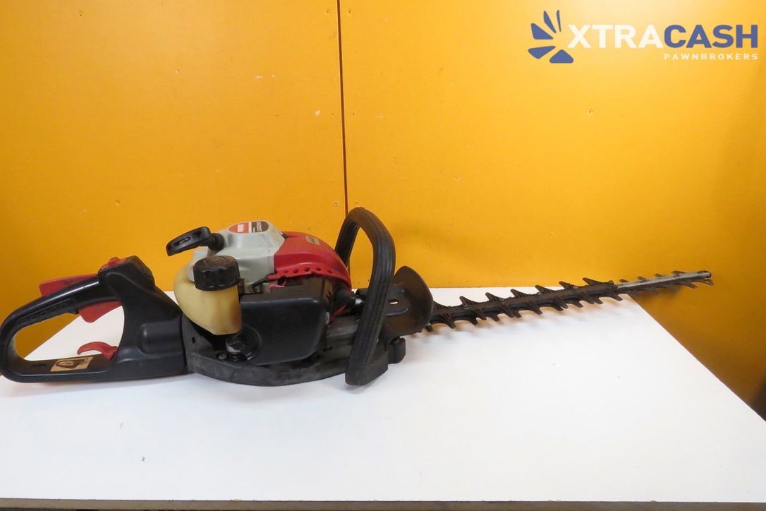 Gas-powered hedge trimmer on a white table against an orange wall with XTRACASH logo.