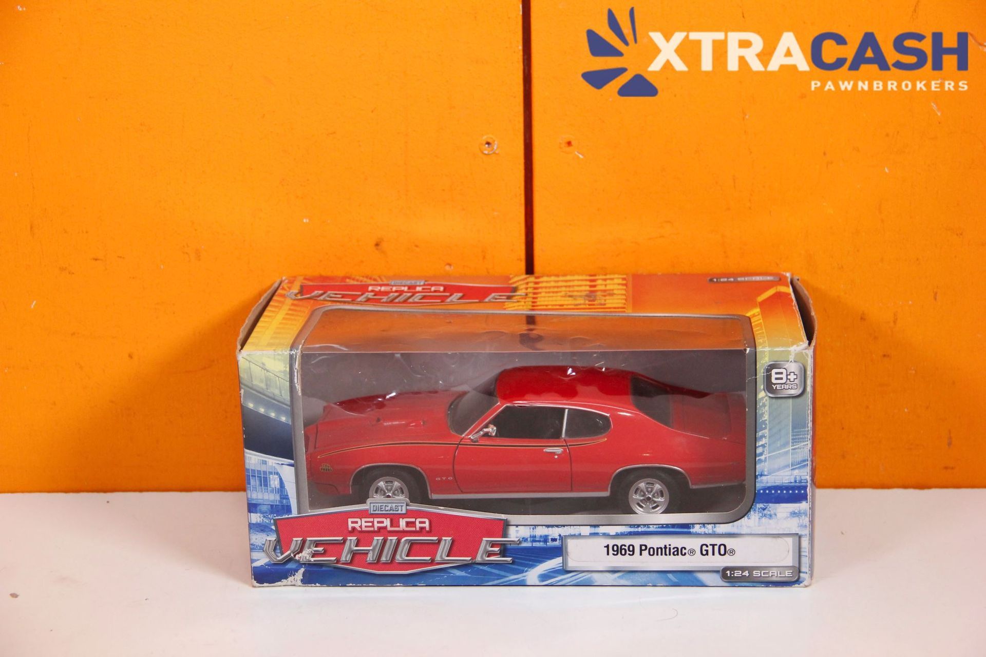Red toy muscle car in a clear box on an orange XtraCash background