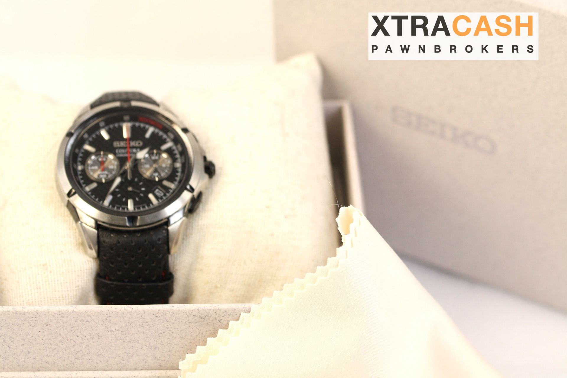Black wristwatch on a white cushion beside an XtraCash Pawn Brokers card