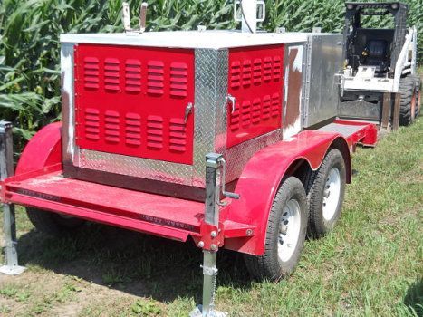 Red equipment trailer with a generator, beside a cornfield and a skid steer.
