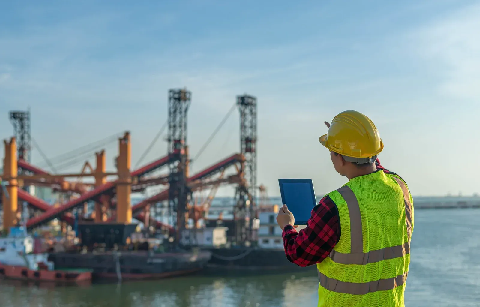 An inspector in a yellow hard hat and reflective vest holds a tablet while overlooking a busy industrial harbor.