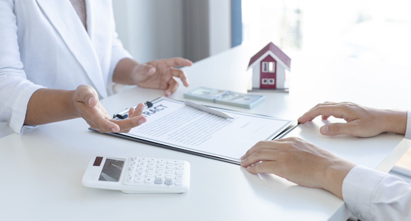 Two people review a contract on a white desk with a small house model, calculator, and cash.