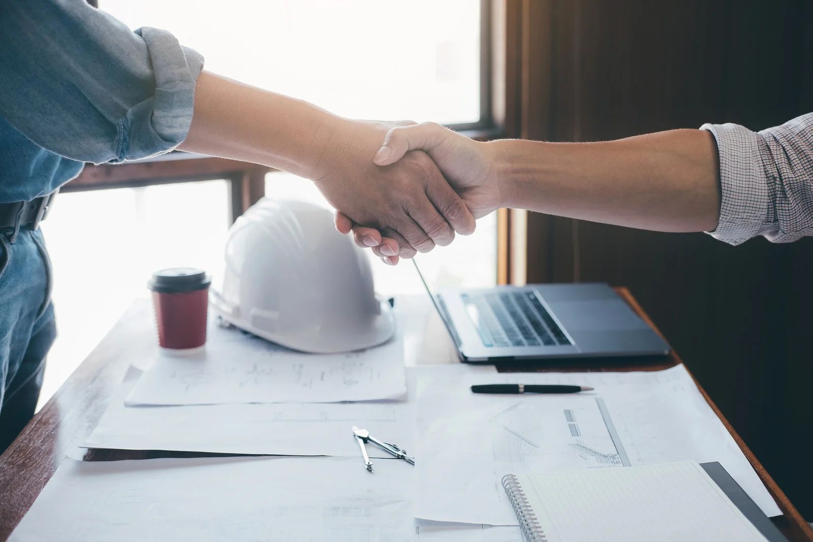 Two people shaking hands across a desk with a laptop, hard hat, and documents