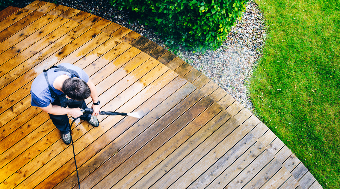 Person pressure washing a wooden deck, cleaning the surface beside a landscaped lawn Person pressure washing a wooden deck, cleaning the surface beside a landscaped lawn