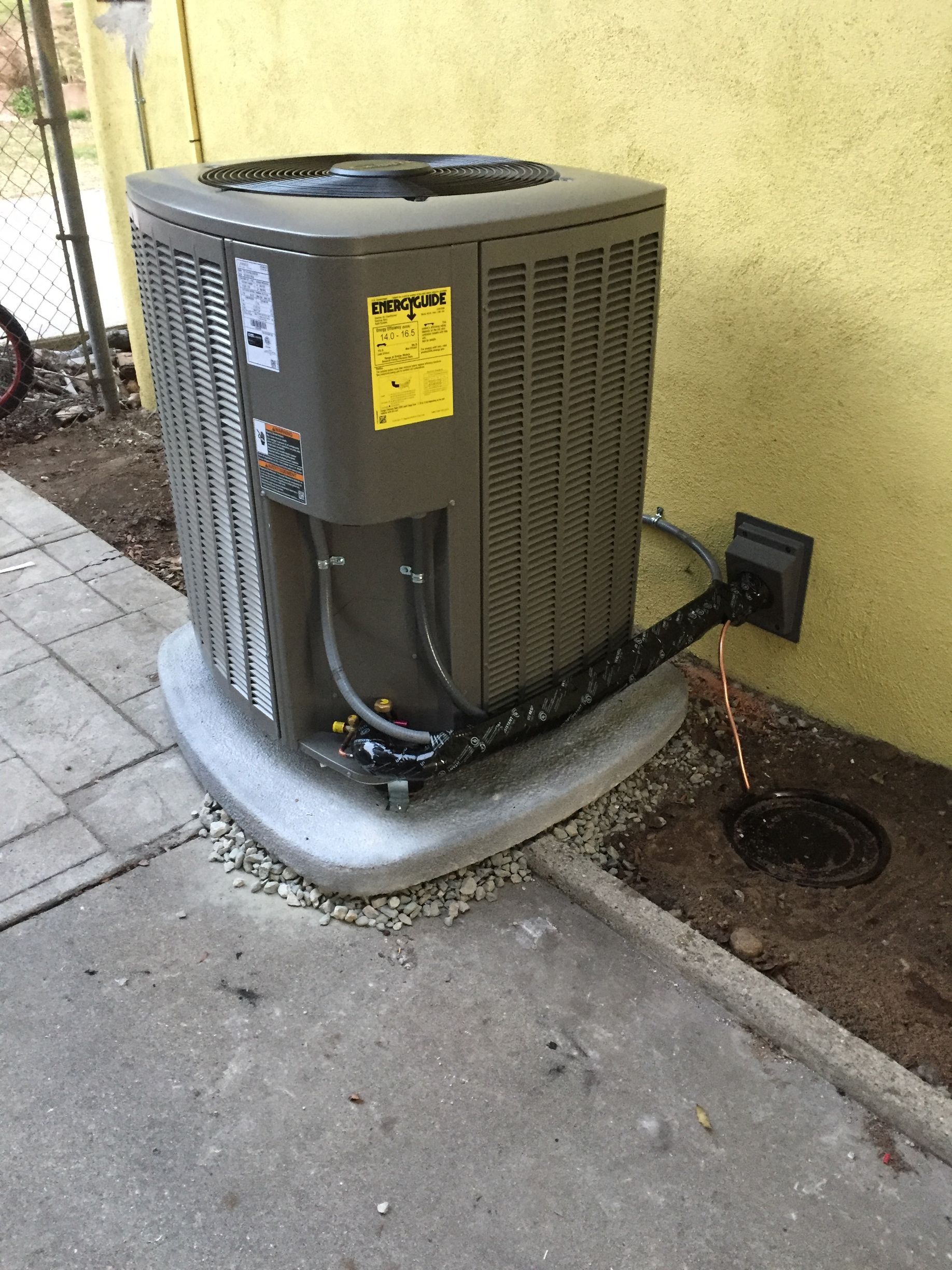 Outdoor HVAC unit beside a building wall on a concrete pad, with a small garden bed nearby