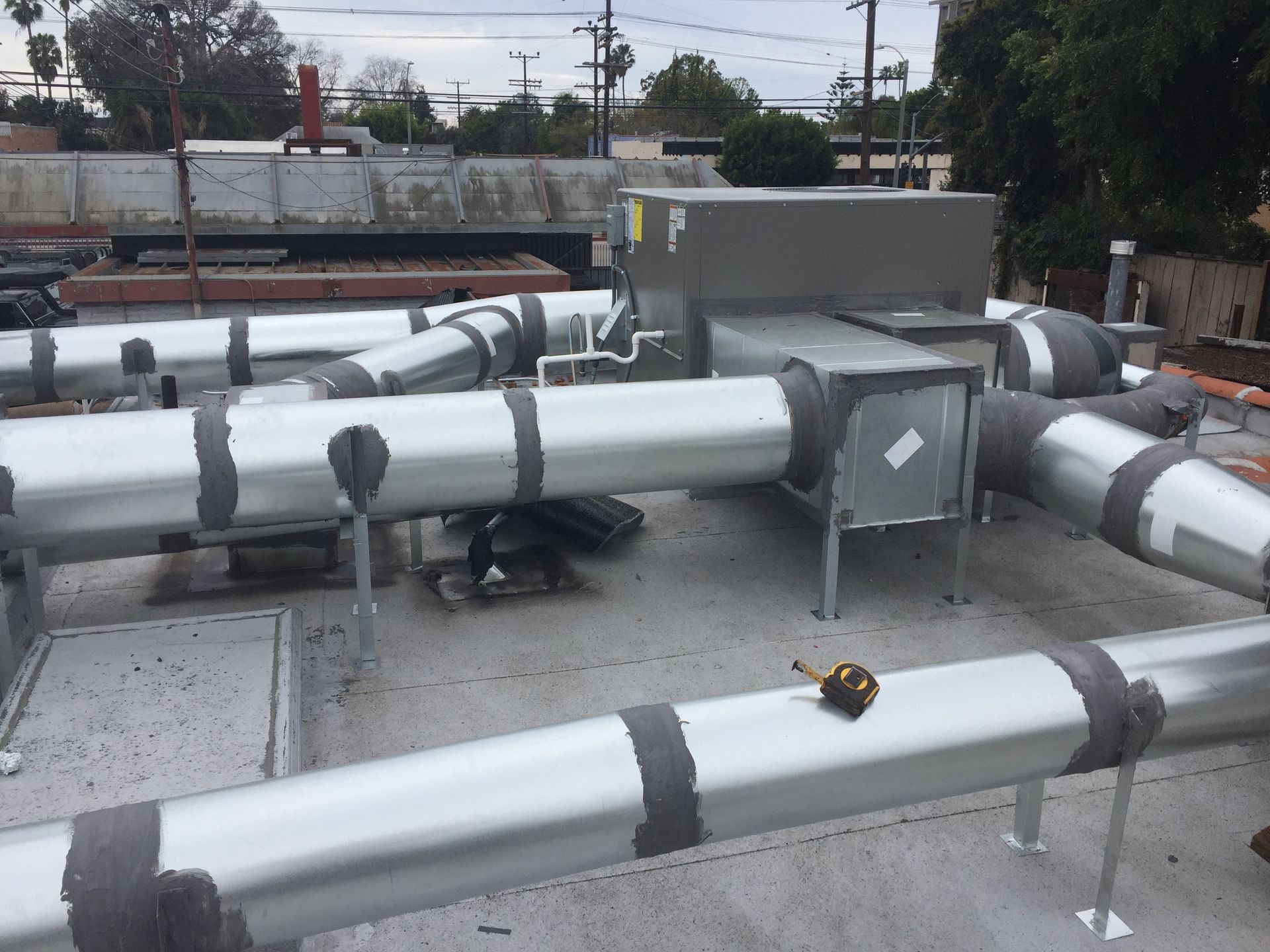 Rooftop HVAC ductwork with silver insulated pipes, gray equipment boxes, and a cloudy sky background.