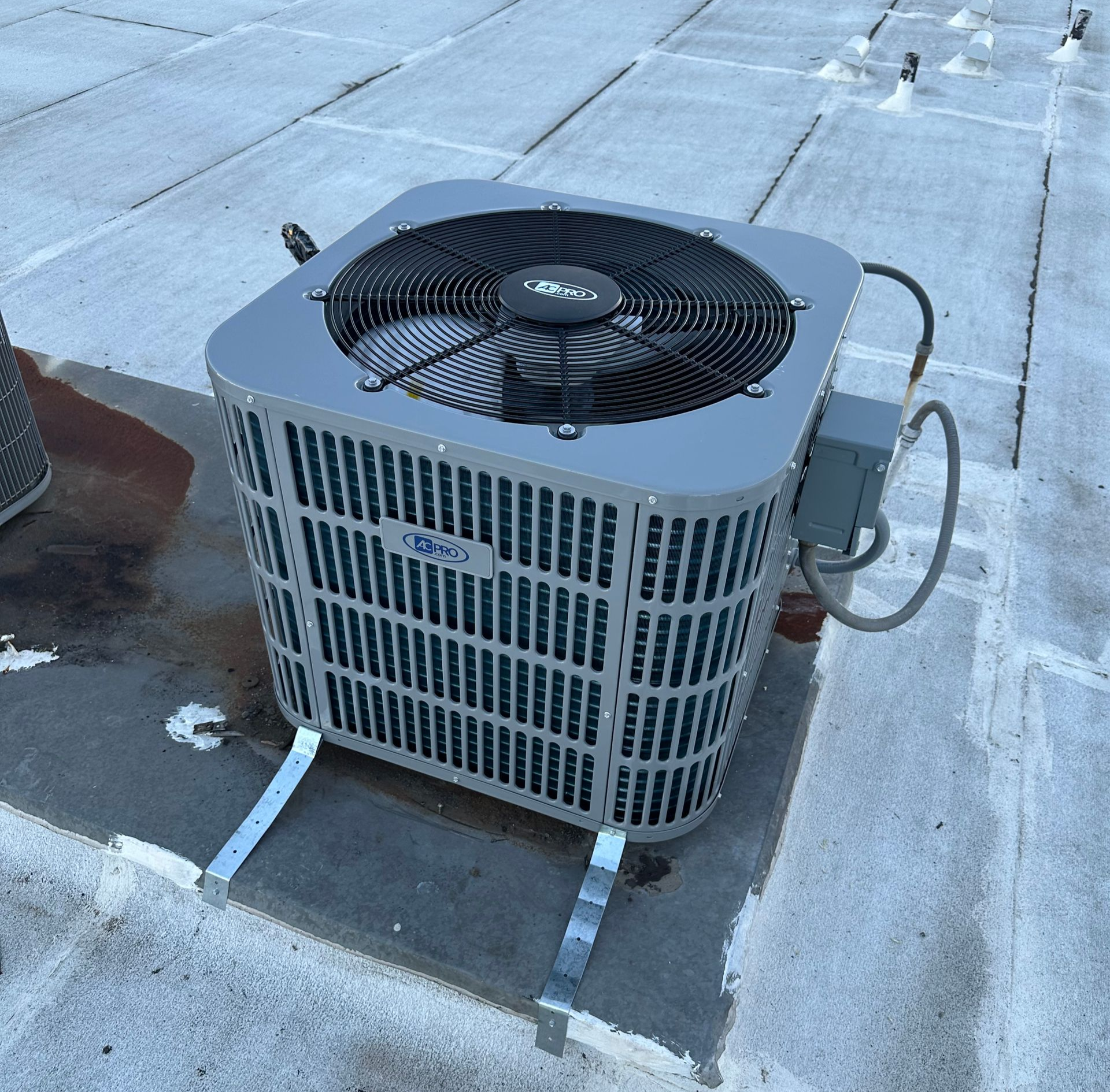 Gray rooftop air conditioner unit on a flat roof with vents and fan visible, connected by a hose