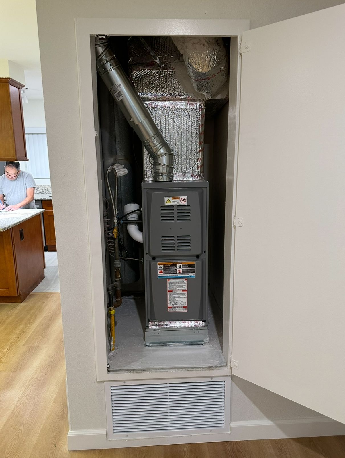 Furnace and ductwork inside a closet. A person is visible in the background in a kitchen.