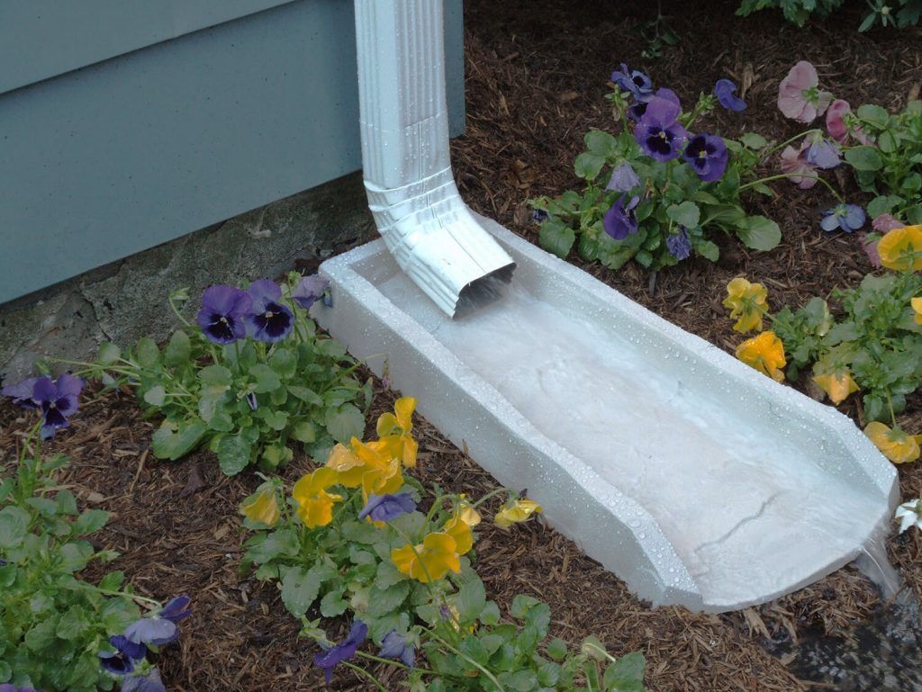 A white gutter downspout drains water into a concrete splash block set in a garden with purple and yellow flowers.
